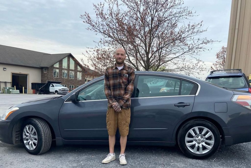 Person standing in front of new-to-them car. They are wearing a plaid shirt and smiling.